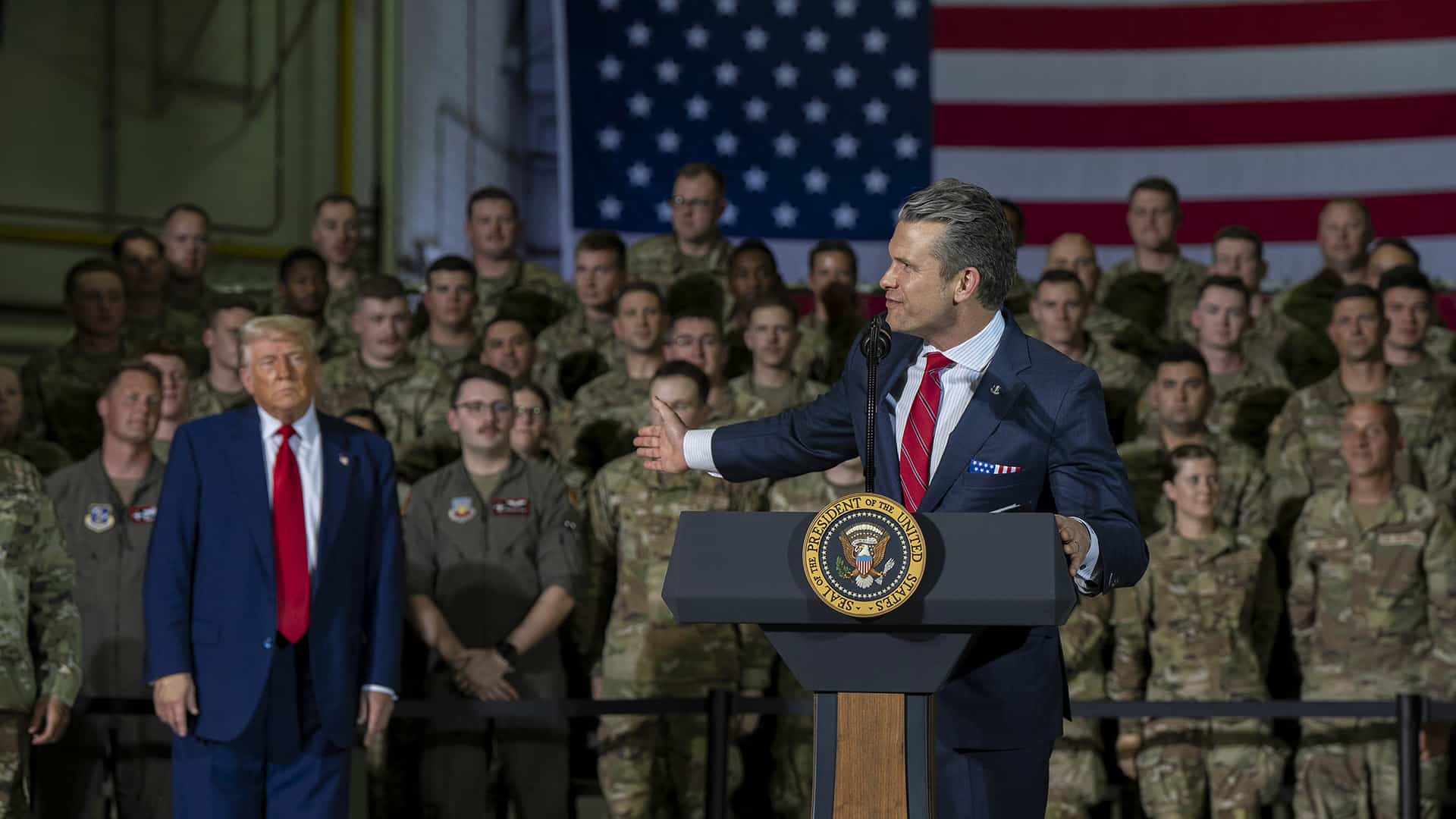 Pete Hegseth pronuncia unas palabras desde un atril con el sello presidencial de Estados Unidos ante el personal militar uniformado y una gran bandera estadounidense en el fondo.