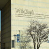 Inscripción “We the People” de la Constitución de Estados Unidos, con fragmentos del texto constitucional, grabada en una fachada de piedra del National Constitution Center en Filadelfia, junto a un árbol sin hojas y farolas con pendones. Al fondo a la izquierda, edificios de ladrillos y la torre de una iglesia con reloj.
