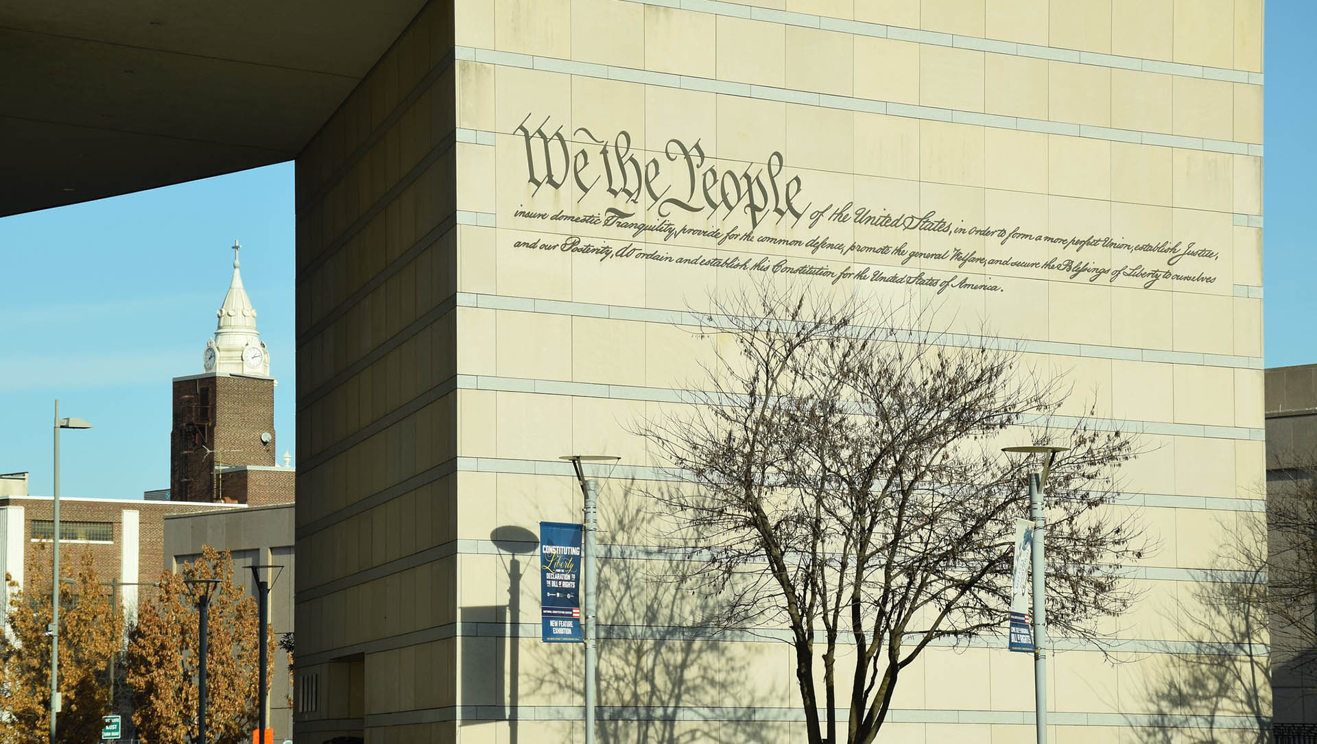Inscripción “We the People” de la Constitución de Estados Unidos, con fragmentos del texto constitucional, grabada en una fachada de piedra del National Constitution Center en Filadelfia, junto a un árbol sin hojas y farolas con pendones. Al fondo a la izquierda, edificios de ladrillos y la torre de una iglesia con reloj.