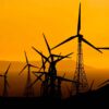 Siluetas de varios aerogeneradores de un parque eólico al atardecer, con sus torres y aspas recortadas sobre un cielo de intenso color naranja. Al fondo se distinguen montañas y el horizonte.