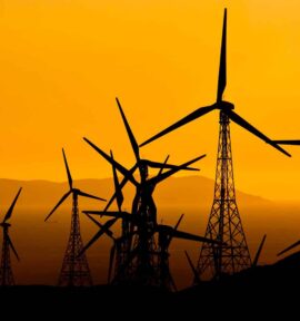 Siluetas de varios aerogeneradores de un parque eólico al atardecer, con sus torres y aspas recortadas sobre un cielo de intenso color naranja. Al fondo se distinguen montañas y el horizonte.