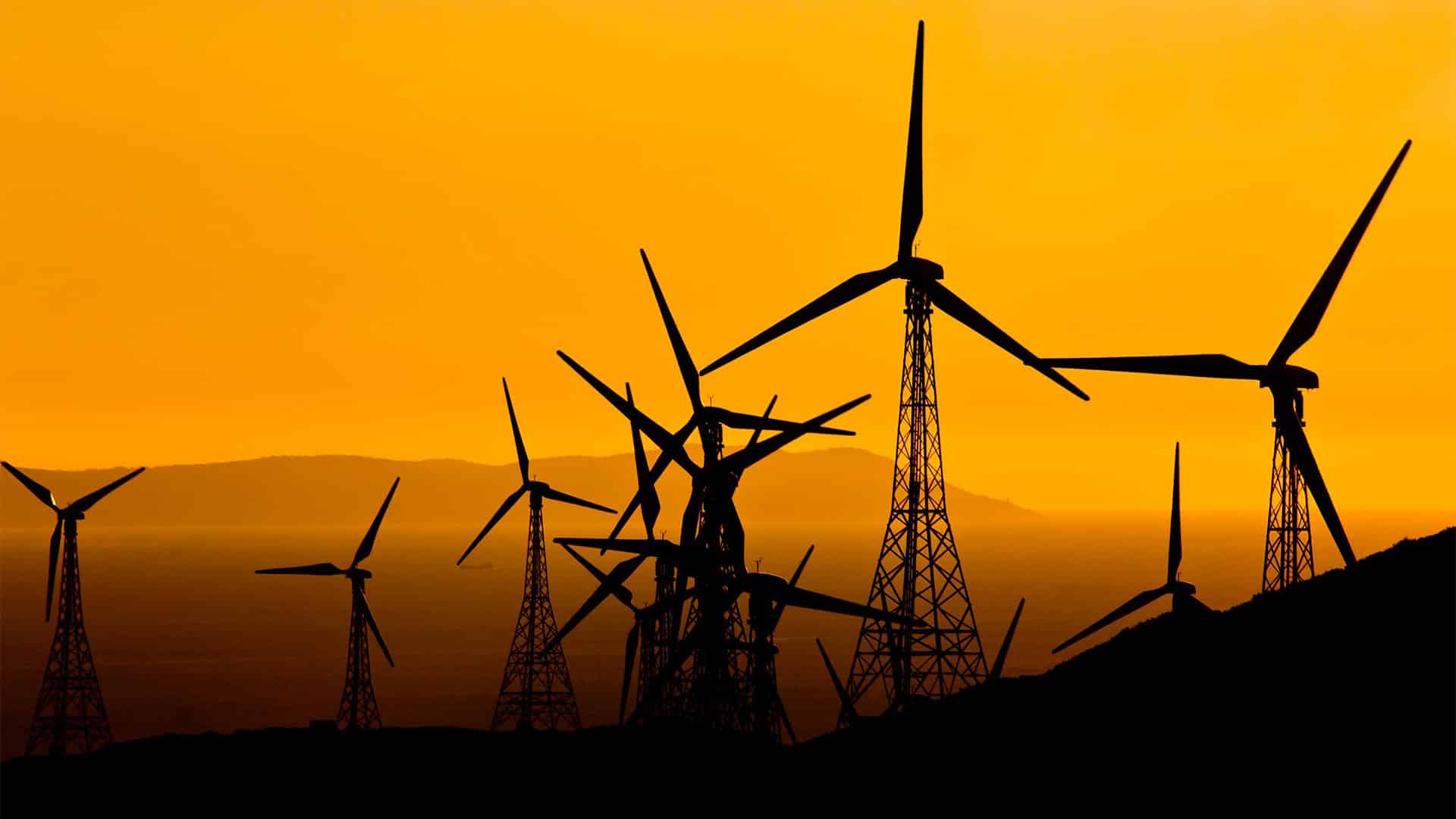 Siluetas de varios aerogeneradores de un parque eólico al atardecer, con sus torres y aspas recortadas sobre un cielo de intenso color naranja. Al fondo se distinguen montañas y el horizonte.